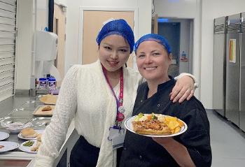 A woman stands with her arm around another woman who is holding a plate of cooked food. Both are wearing hairnets and smiling to camera.