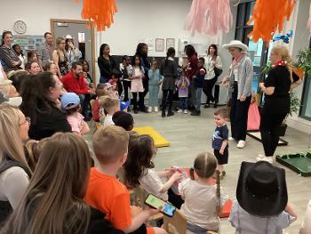 A room full of children gathered around to listen to an adult wearing a cowboy hat.