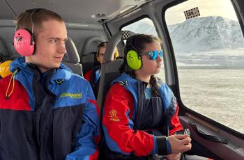 Two pupils look out the window of a helicopter landing near a snowy hill.