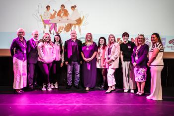 Four young people and seven adults stand on a stage in a row under purple lighting.