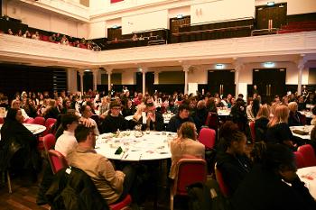 Hundreds of young people sit at round tables in a large room.