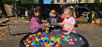 Three young children play with coloured block on a table outside in the sunshine.