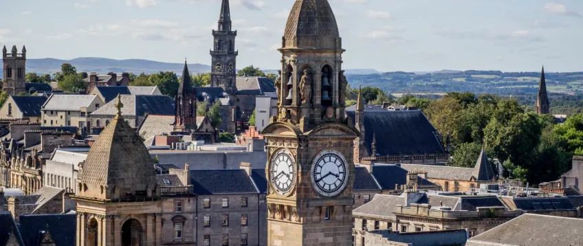Scenic view of Paisley town centre in Renfrewshire, with the historic clock tower of Paisley Town Hall in the foreground, Victorian and Gothic buildings around it, church spires, and green hills in the distance under a partly cloudy sky.