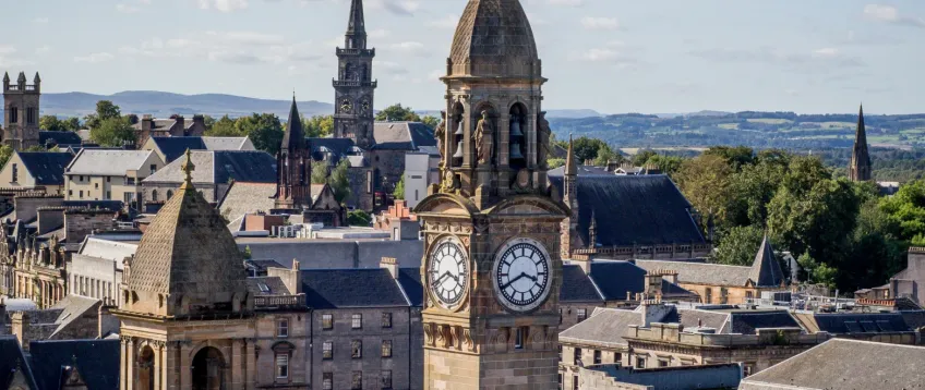 Scenic view of Paisley town centre in Renfrewshire, with the historic clock tower of Paisley Town Hall in the foreground, Victorian and Gothic buildings around it, church spires, and green hills in the distance under a partly cloudy sky.