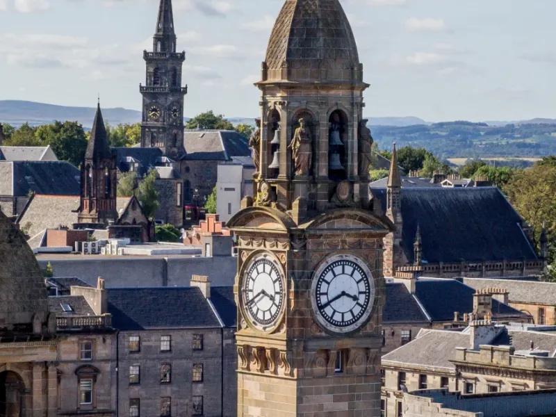 Scenic view of Paisley town centre in Renfrewshire, with the historic clock tower of Paisley Town Hall in the foreground, Victorian and Gothic buildings around it, church spires, and green hills in the distance under a partly cloudy sky.