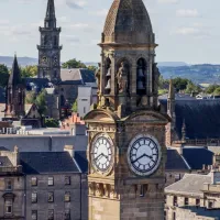 Scenic view of Paisley town centre in Renfrewshire, with the historic clock tower of Paisley Town Hall in the foreground, Victorian and Gothic buildings around it, church spires, and green hills in the distance under a partly cloudy sky.