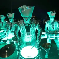 Five performers in shiny, light-up costumes are playing drums at night. A crowd is watching in the background.