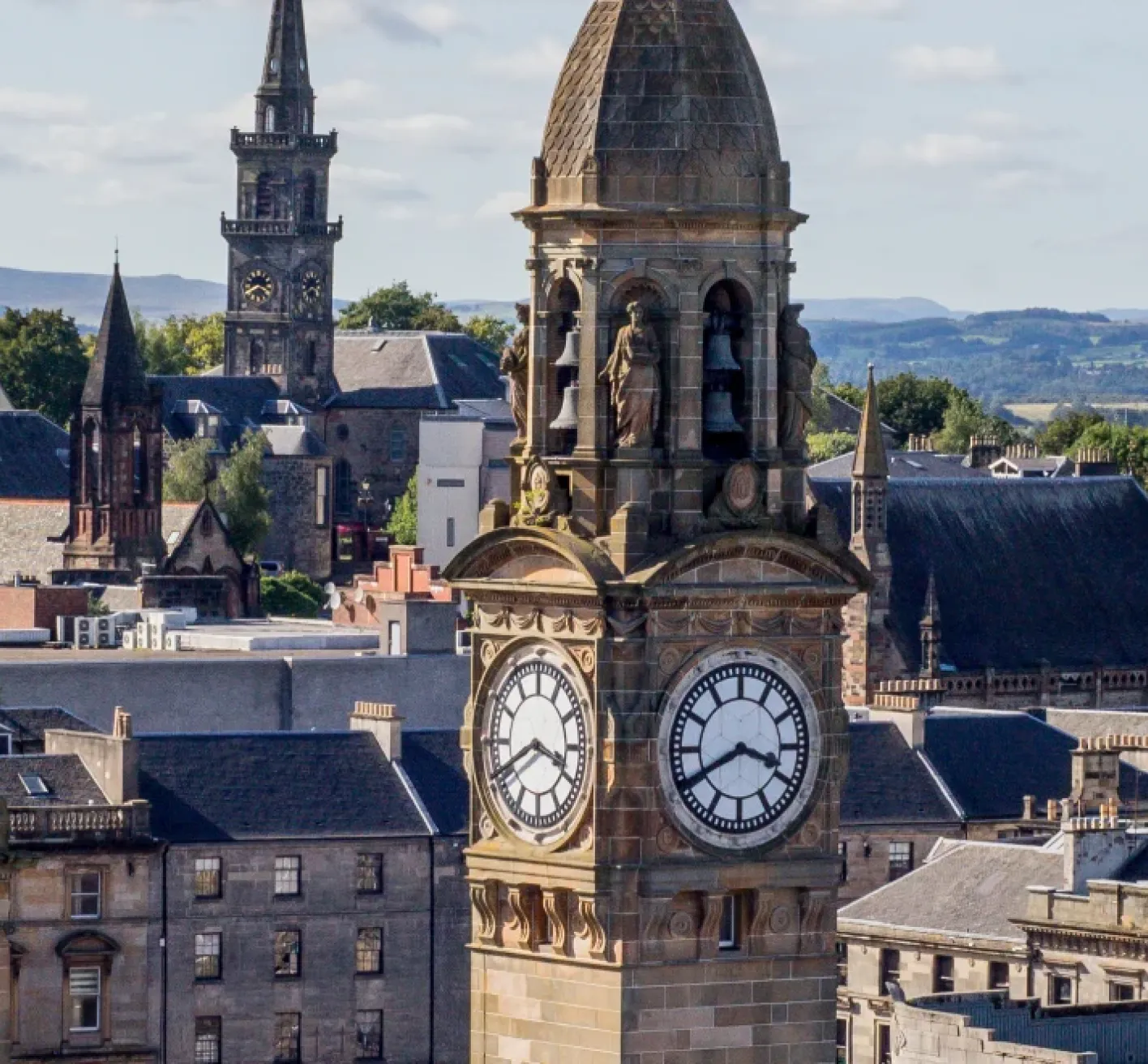 Scenic view of Paisley town centre in Renfrewshire, with the historic clock tower of Paisley Town Hall in the foreground, Victorian and Gothic buildings around it, church spires, and green hills in the distance under a partly cloudy sky.