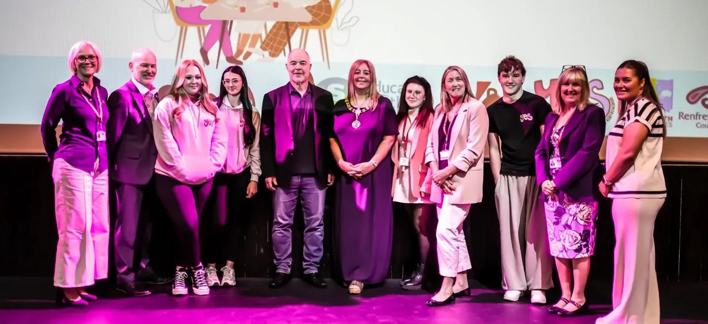 11 people standing and smiling on a purple stage