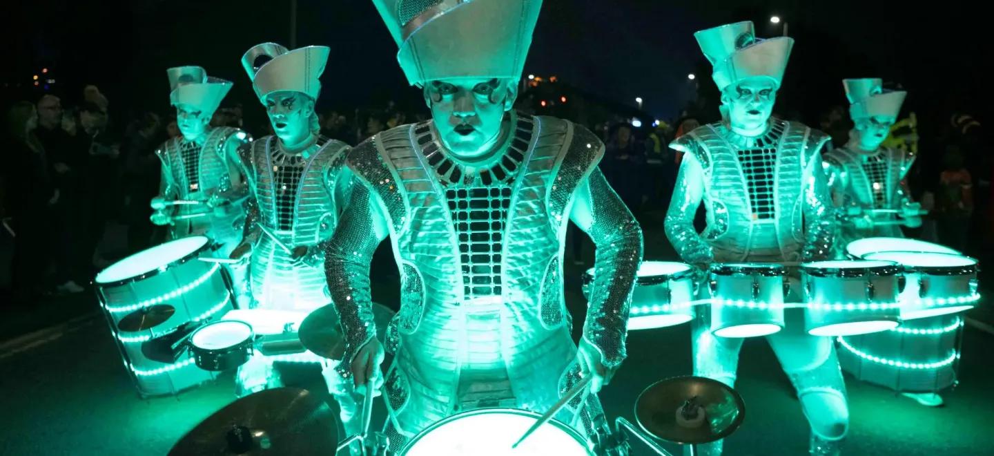 Five performers in shiny, light-up costumes are playing drums at night. A crowd is watching in the background.