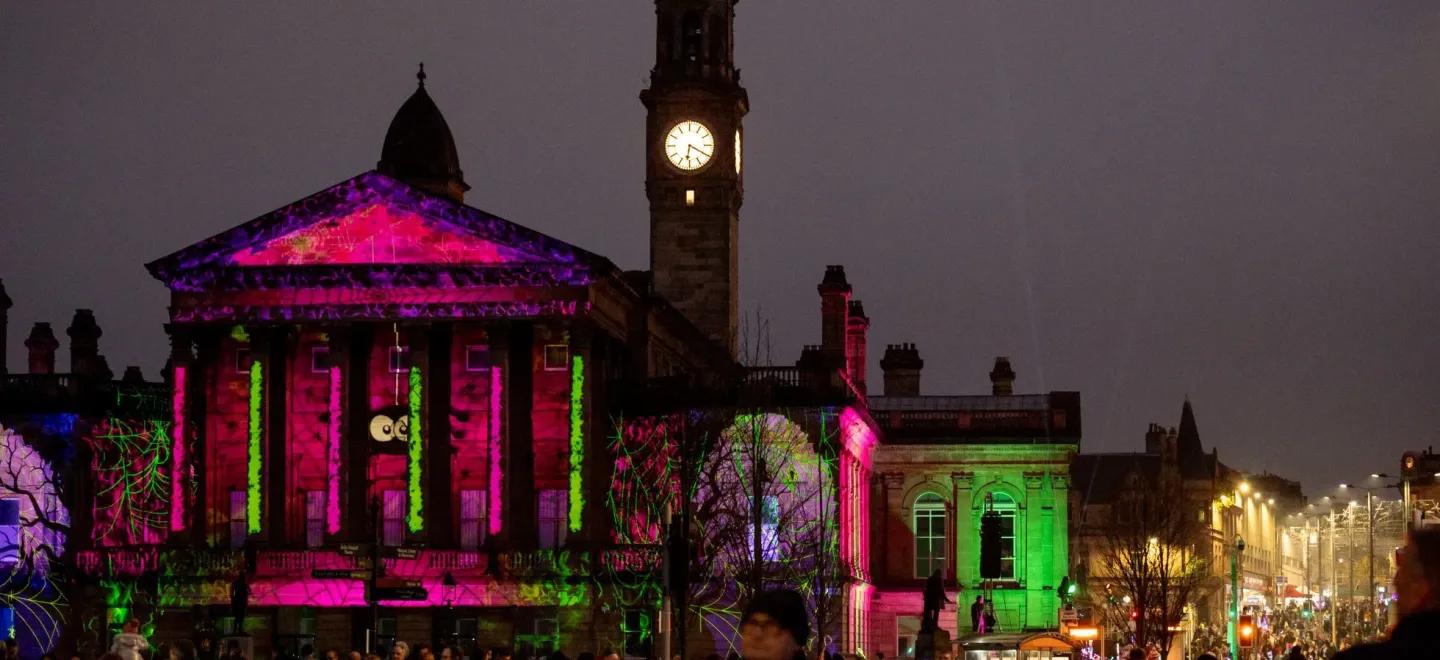 Crowds at Paisley Halloween Parade in front of a lit-up historic building with spooky projections.