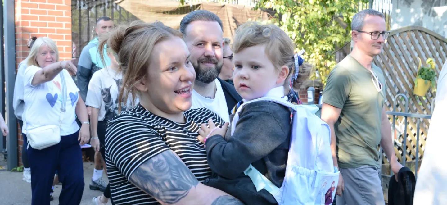 A parent holding a child and walking into a school playground with other parents and children.