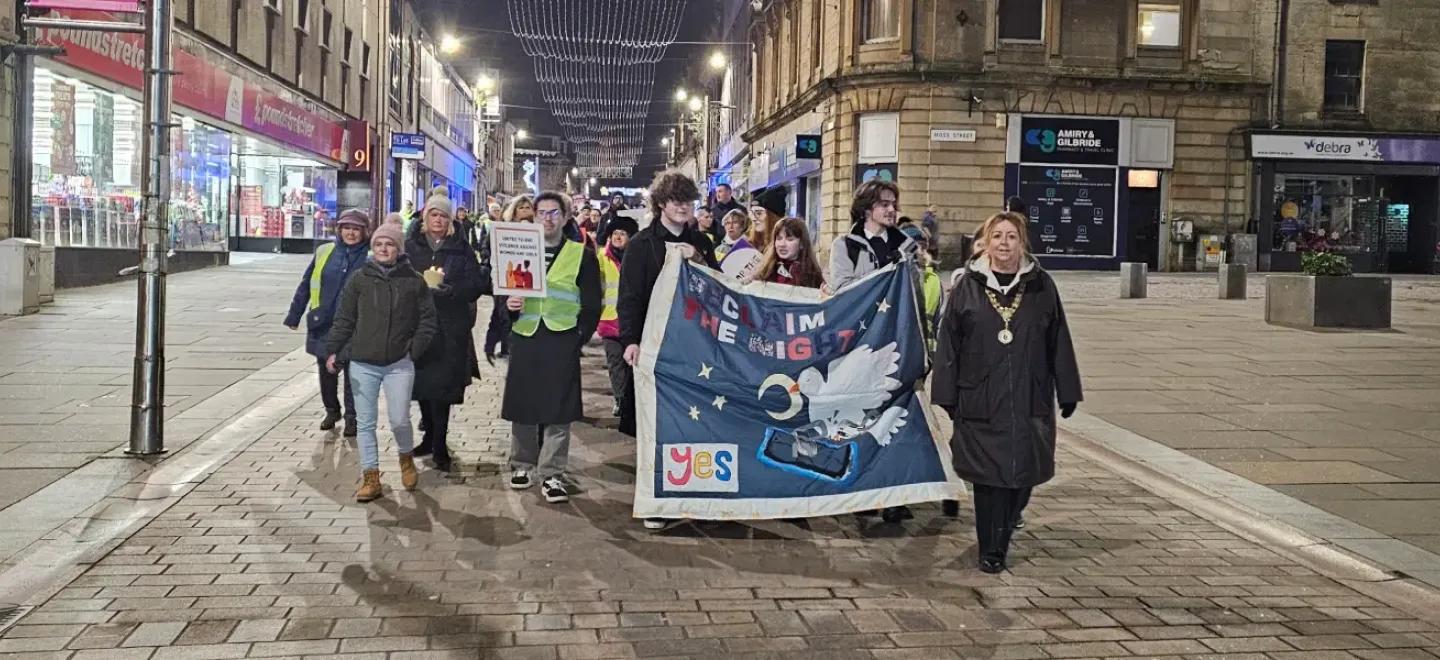 A large group of people walk down an urban street at night behind a banner proclaiming, "Reclaim the Night."