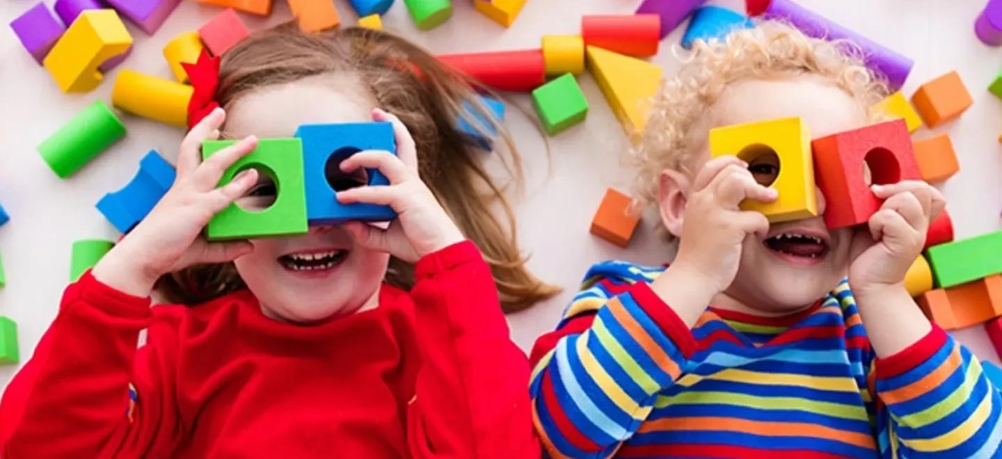Two young children lying on their backs surrounded by colourful block and holding two over their eyes like glasses.