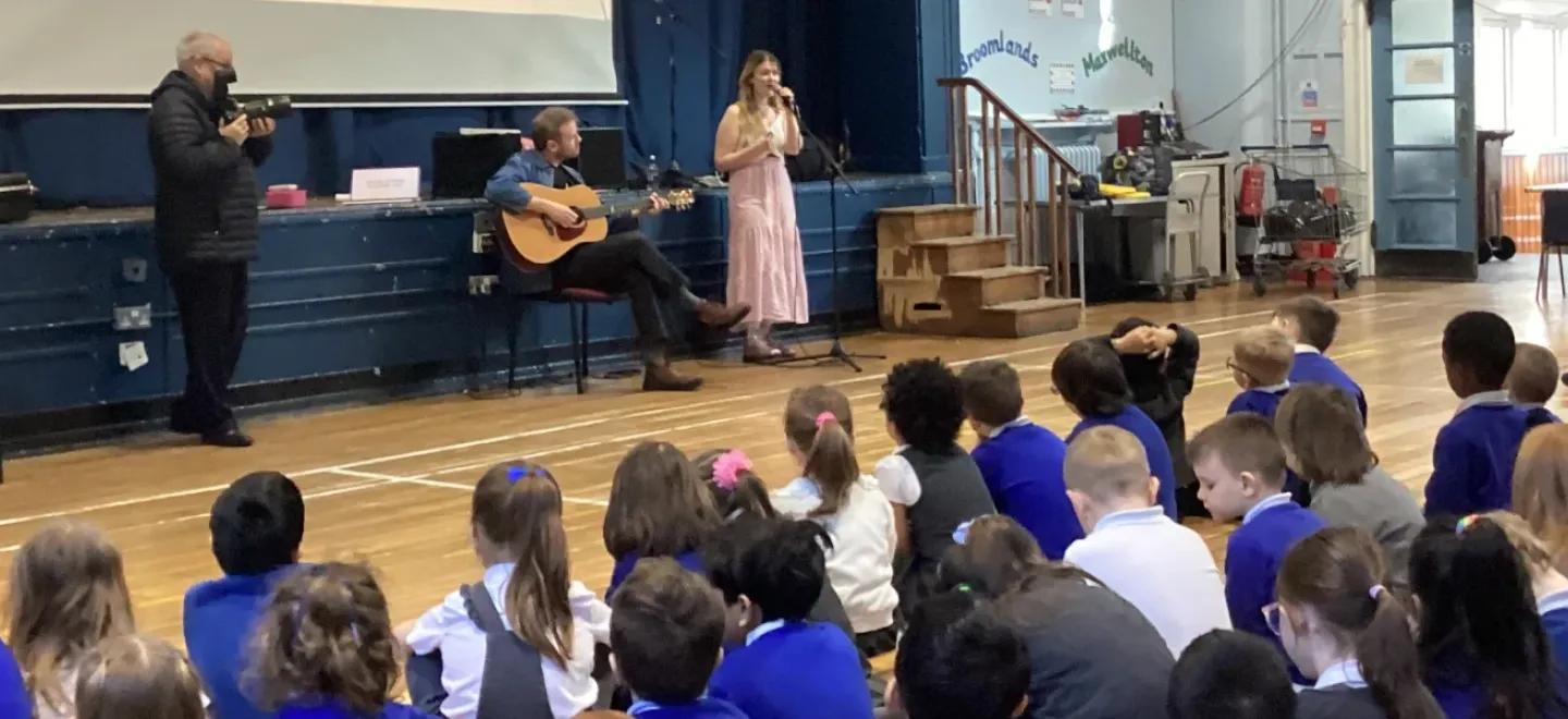 Pupils sitting in a hall listening to a signer and guitarist.