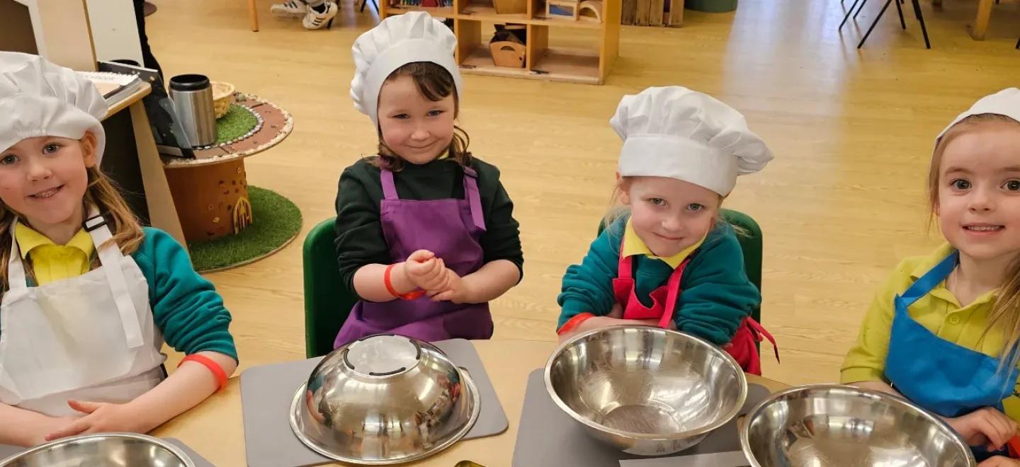 Four young children wear chef's hats and sit at a table with baking equipment laid out.