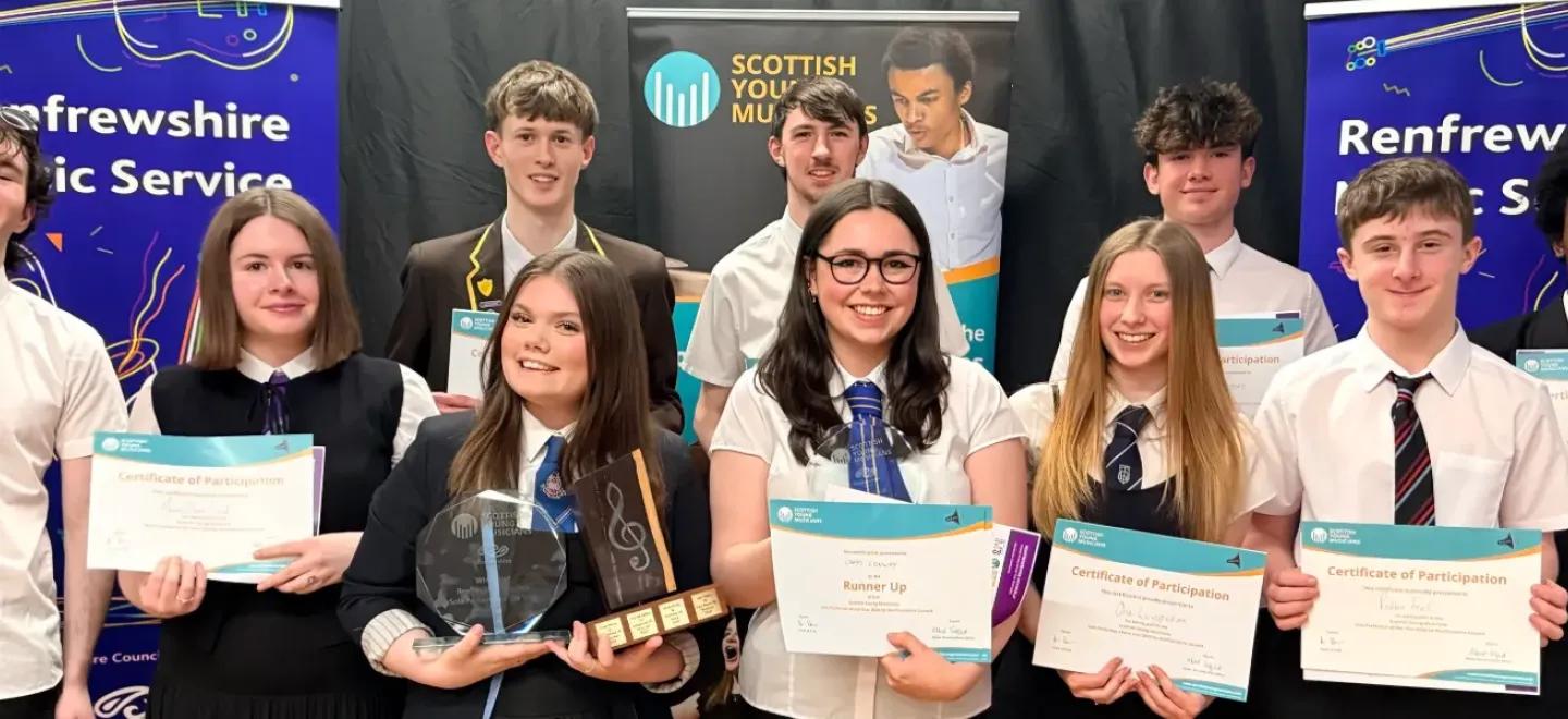 Ten young people stand together smiling and holding their certificates and awards.