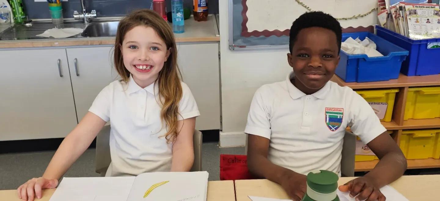 Two pupils smile, seated in their classroom to write in their jotters.