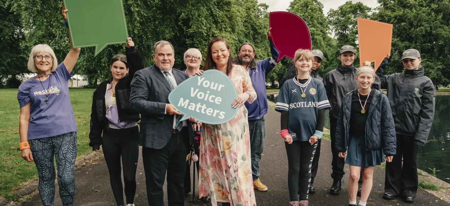 11 people standing in a park holding Your Voice Matters colourful signs