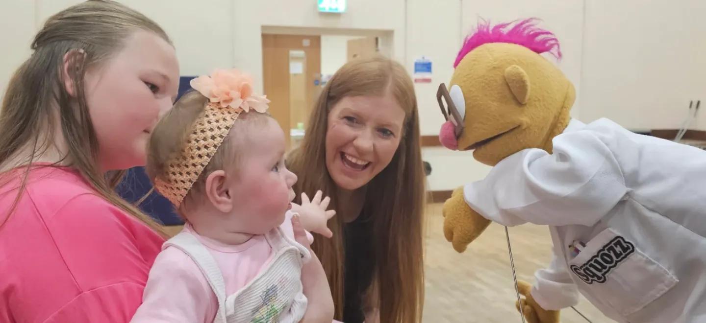 A young child interacts with a puppet while being held by relatives.