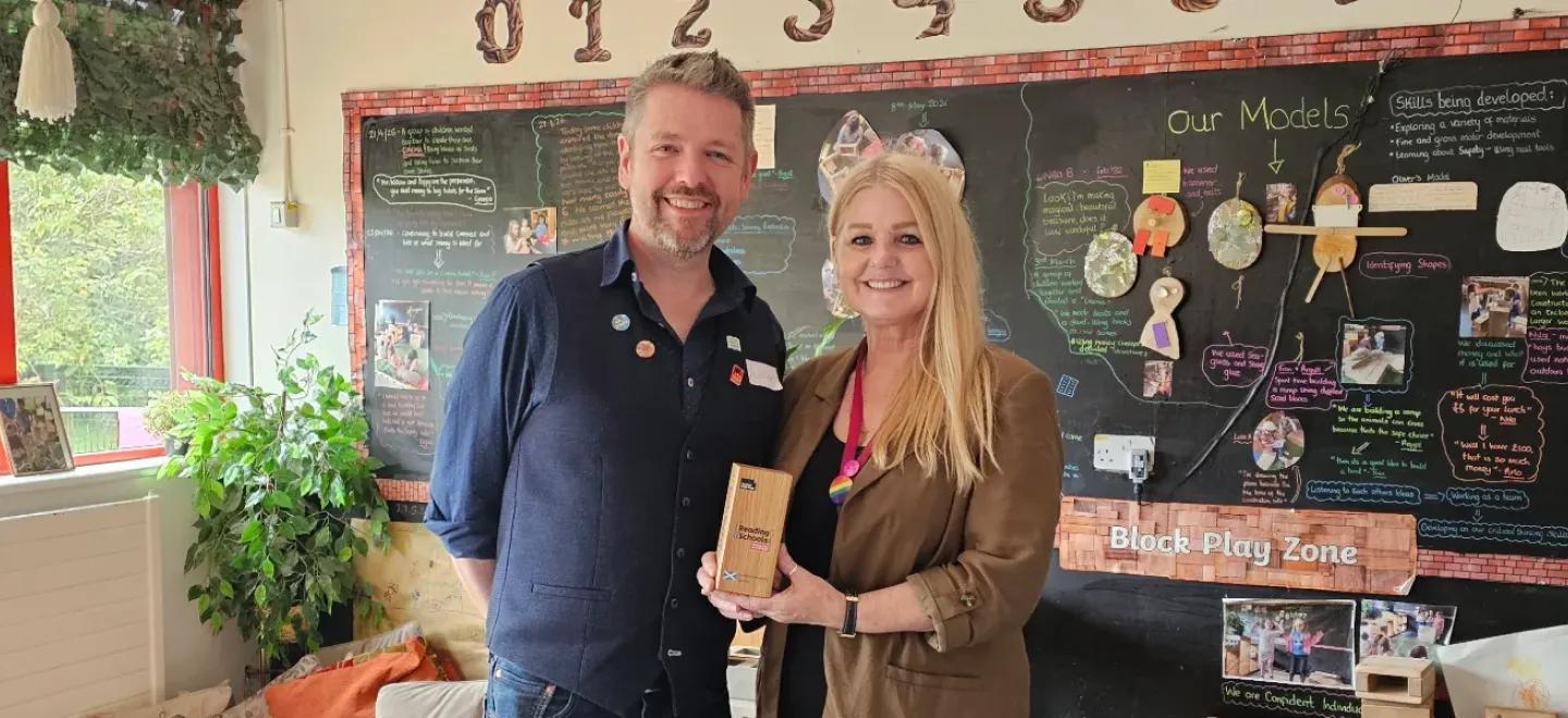 Two adults stand in a nursery and smile. One holds a trophy.