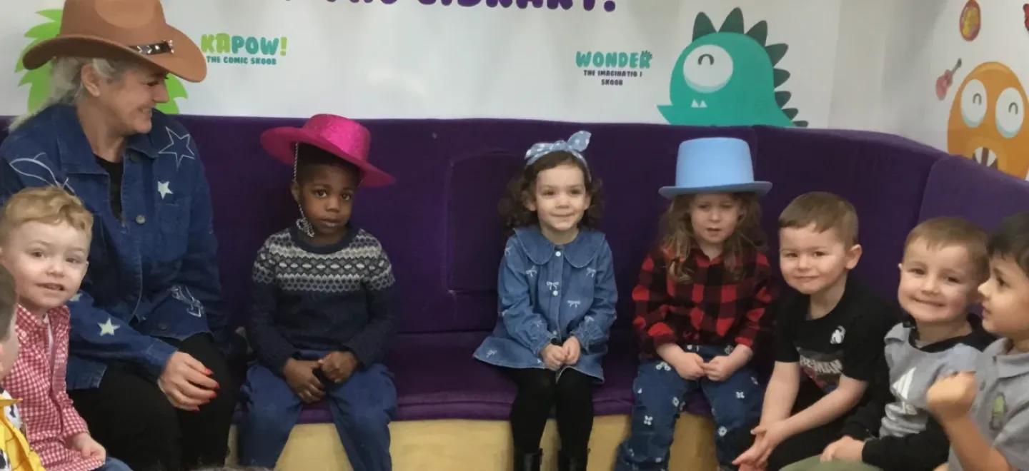 Children sit on a corner reading seat with adults. A few are wearing cowboy hats. A cardboard cut out of Dolly Parton is at the left-hand side.
