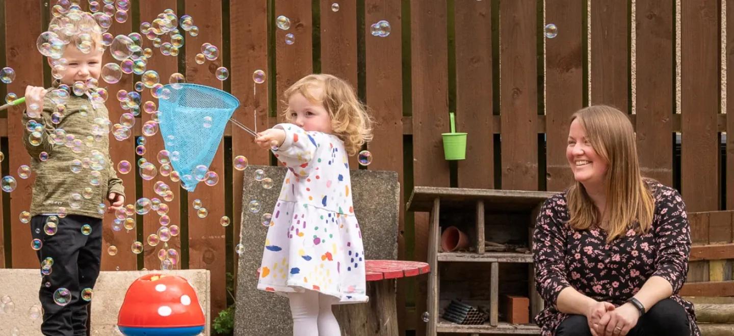 Two children play with bubbles in a garden supervised by an adult.
