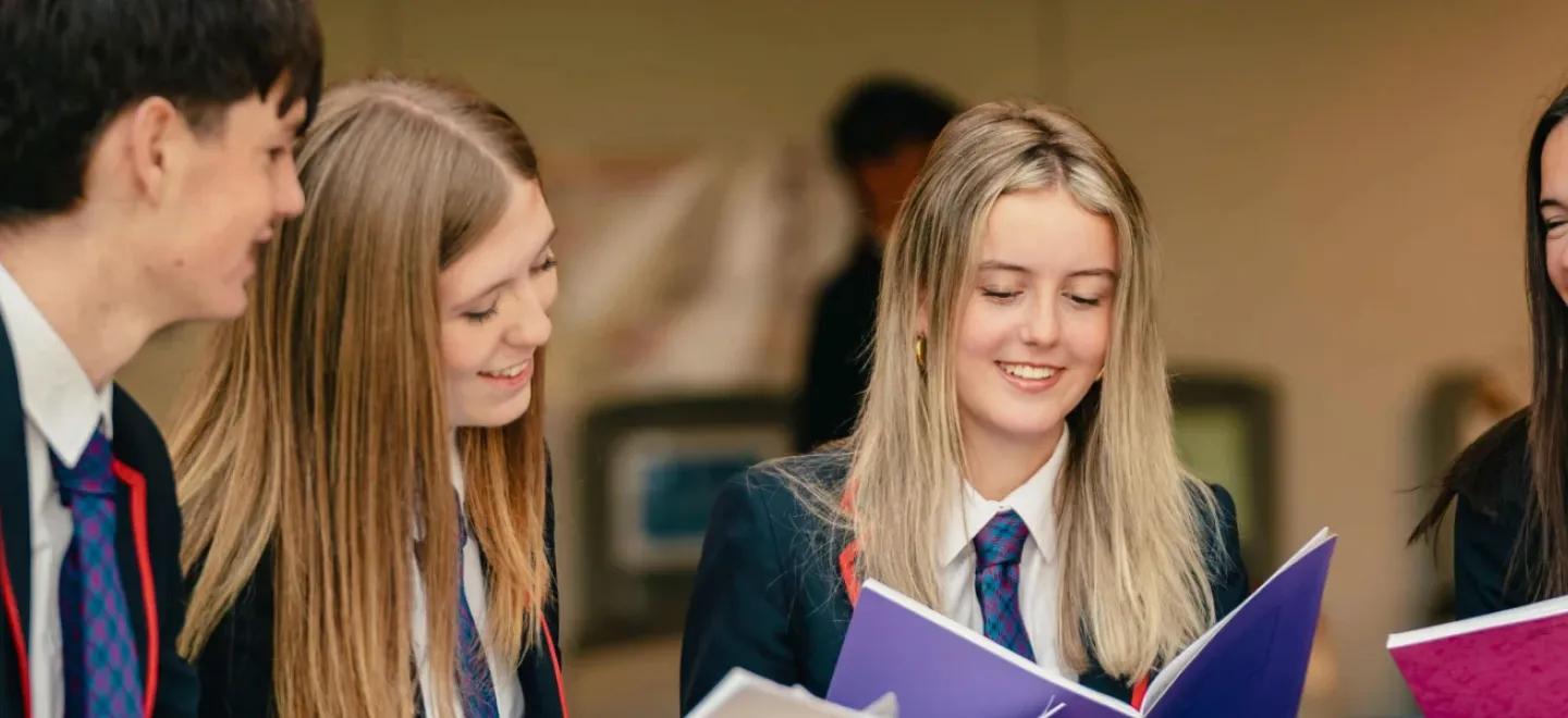 Four high school pupils smile as they hold notebooks and discuss coursework.