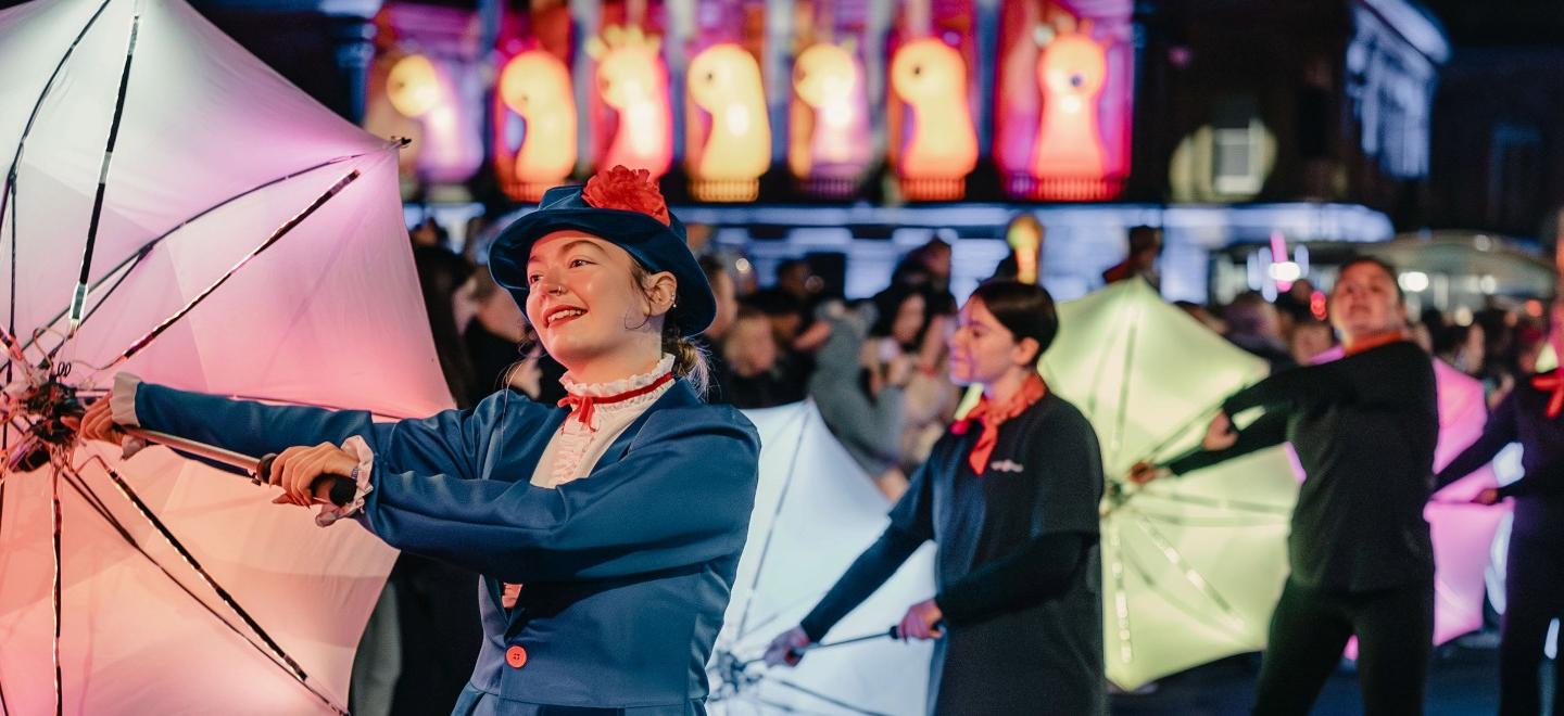 Performers carrying illuminated umbrellas during the Paisley Halloween Festival parade, with colourful light projections and illuminated installations visible in the background.