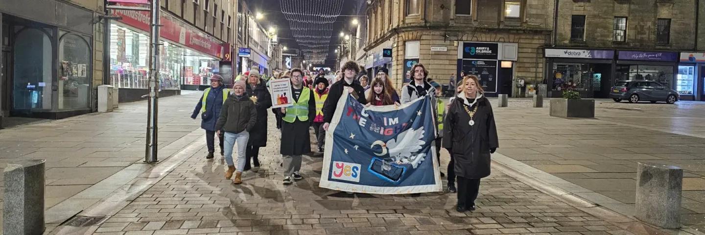 Young people, adults and Renfrewshire's Provost march down an urban street with a Reclaim the Night banner.