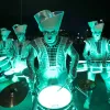 Five performers in shiny, light-up costumes are playing drums at night. They’re wearing big, angular hats and glowing outfits. A crowd is watching in the background.